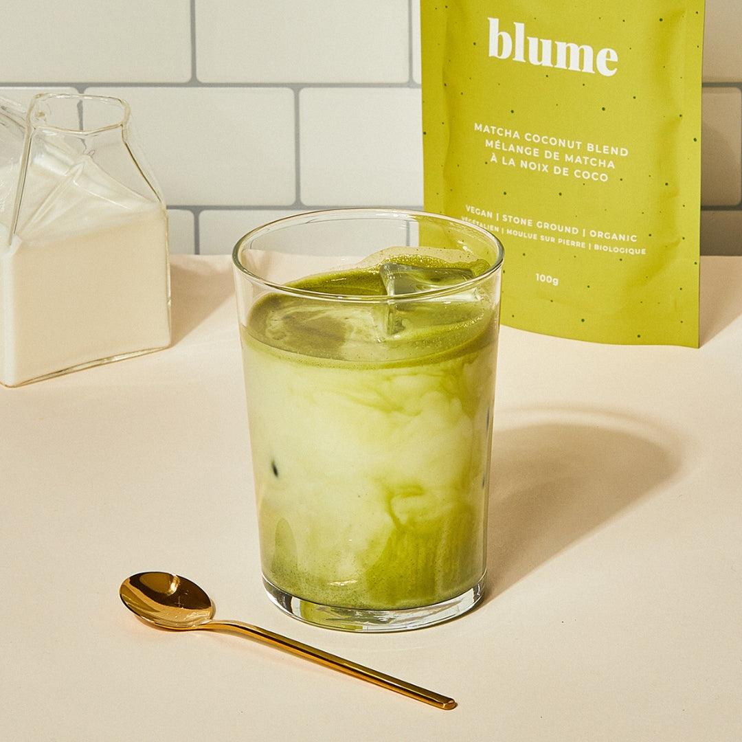 A glass of matcha latte with swirling green and white layers sits on a light surface. A small gold spoon is nearby. Behind the glass is a partially visible milk container and a yellow package labeled blume with matcha coconut blend details.